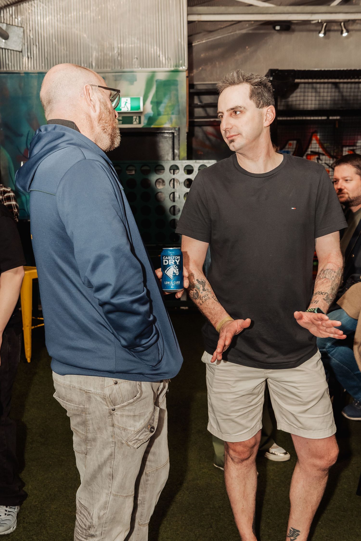 Two tattooed men conversing at an indoor event, one holding a can of beer, with industrial decor in the background.