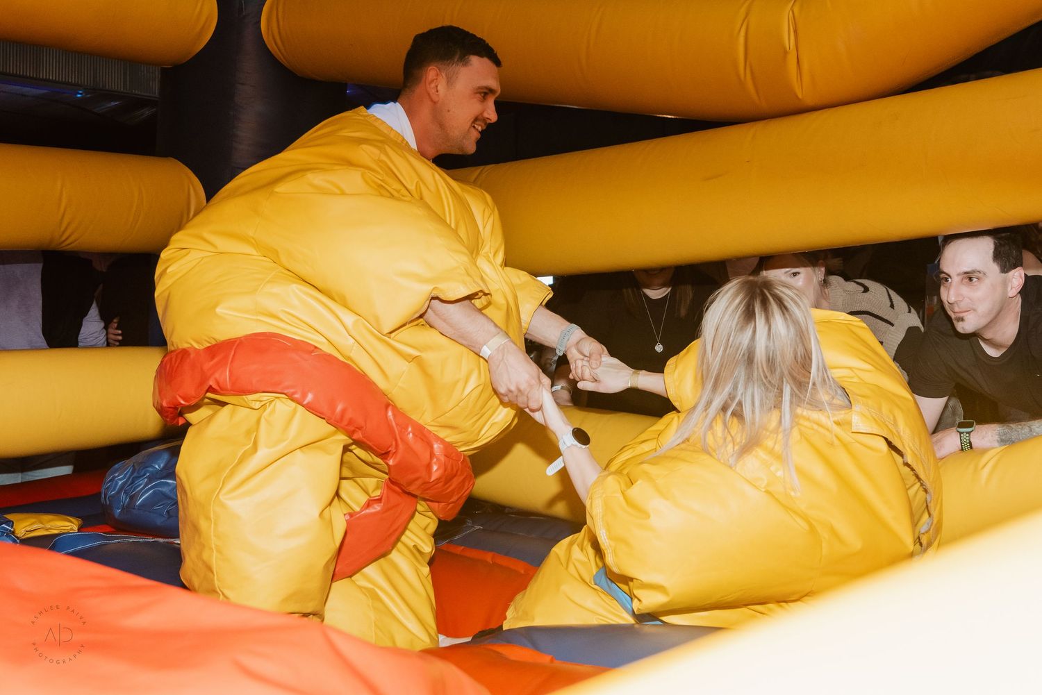Two people in yellow sumo wrestling suits compete in an inflatable bouncy arena at a fun event.