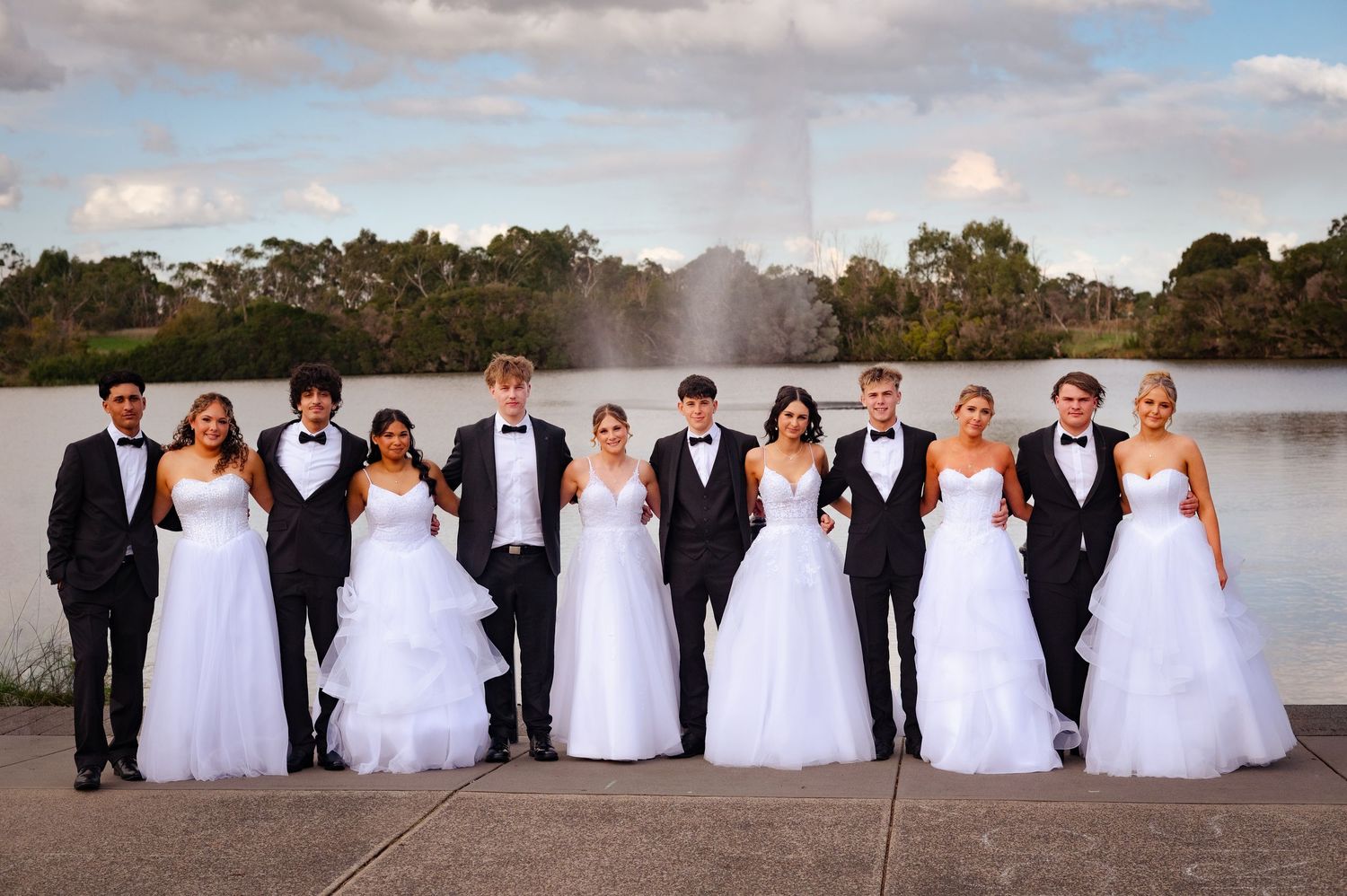 Group of elegantly dressed teens in black tuxedos and white gowns posing by a lake with a fountain at prom.