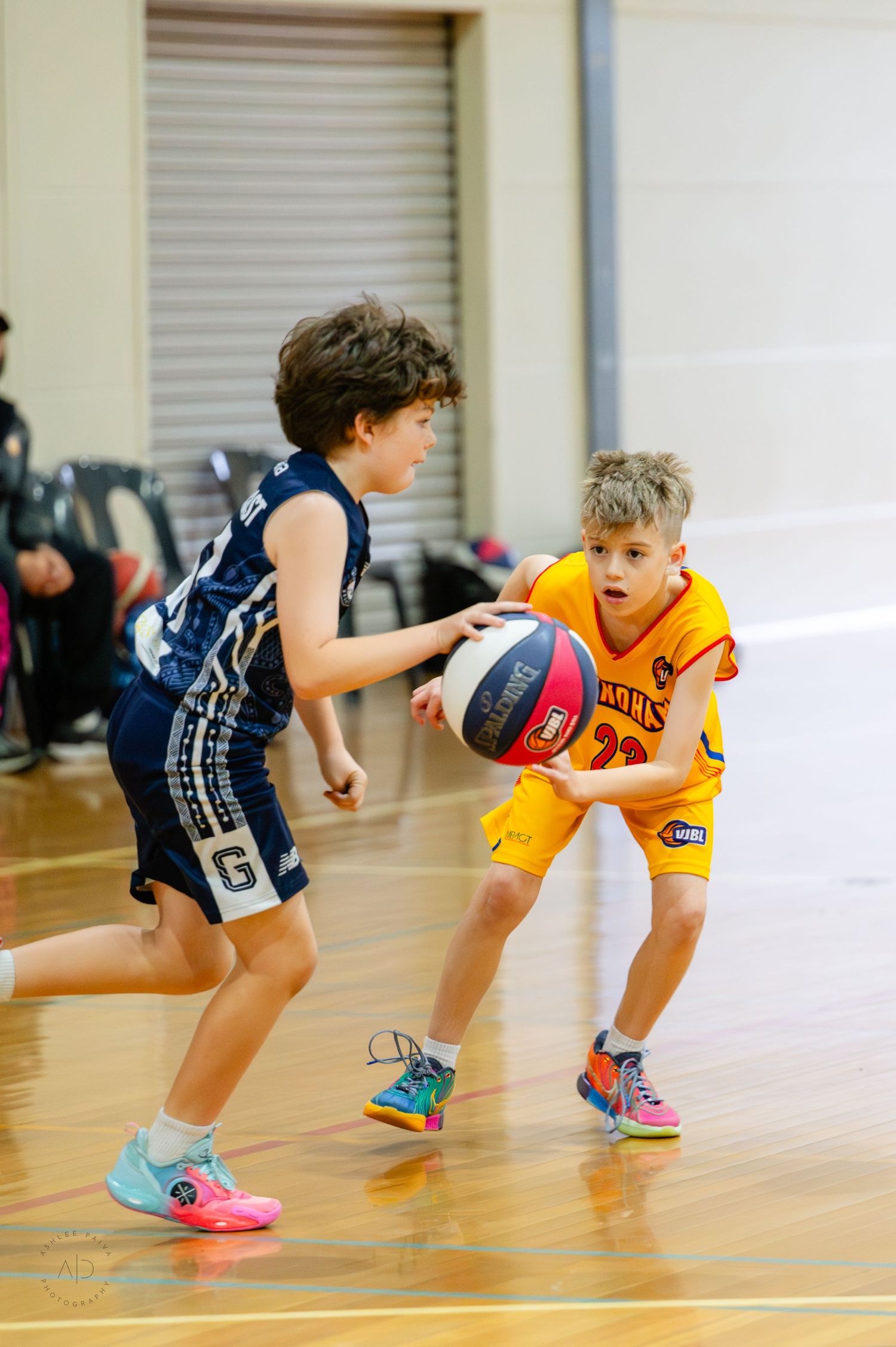 Two young boys playing basketball in a gym, one in navy uniform dribbling against a defender in yellow.