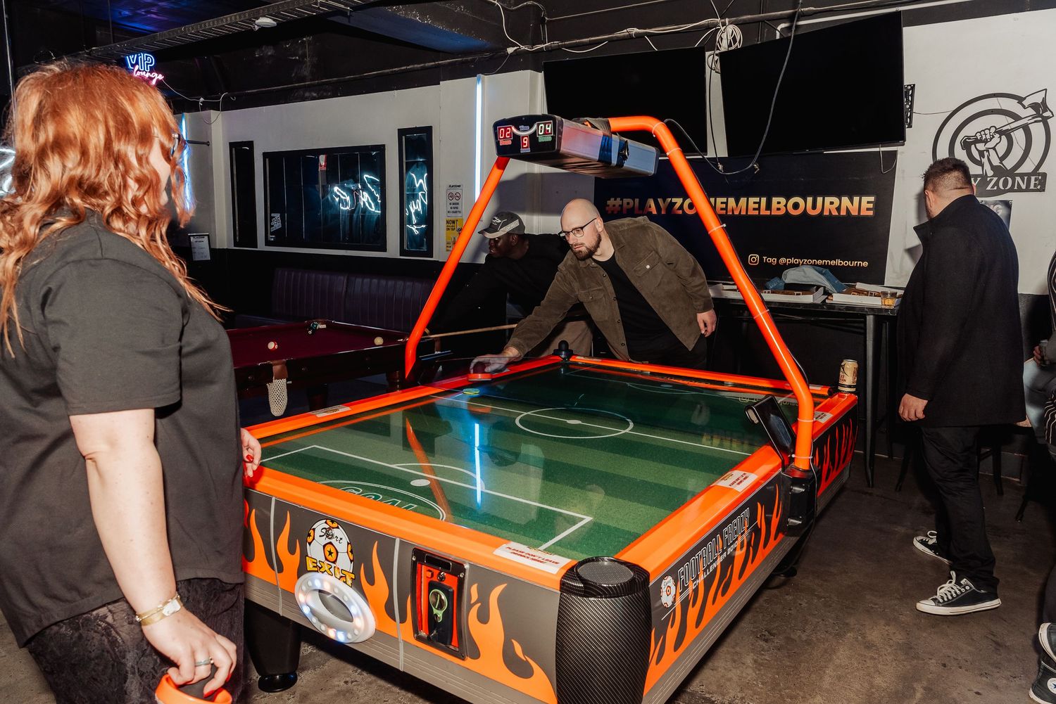 People playing air hockey on an orange flame-decorated table in a dark arcade game room.