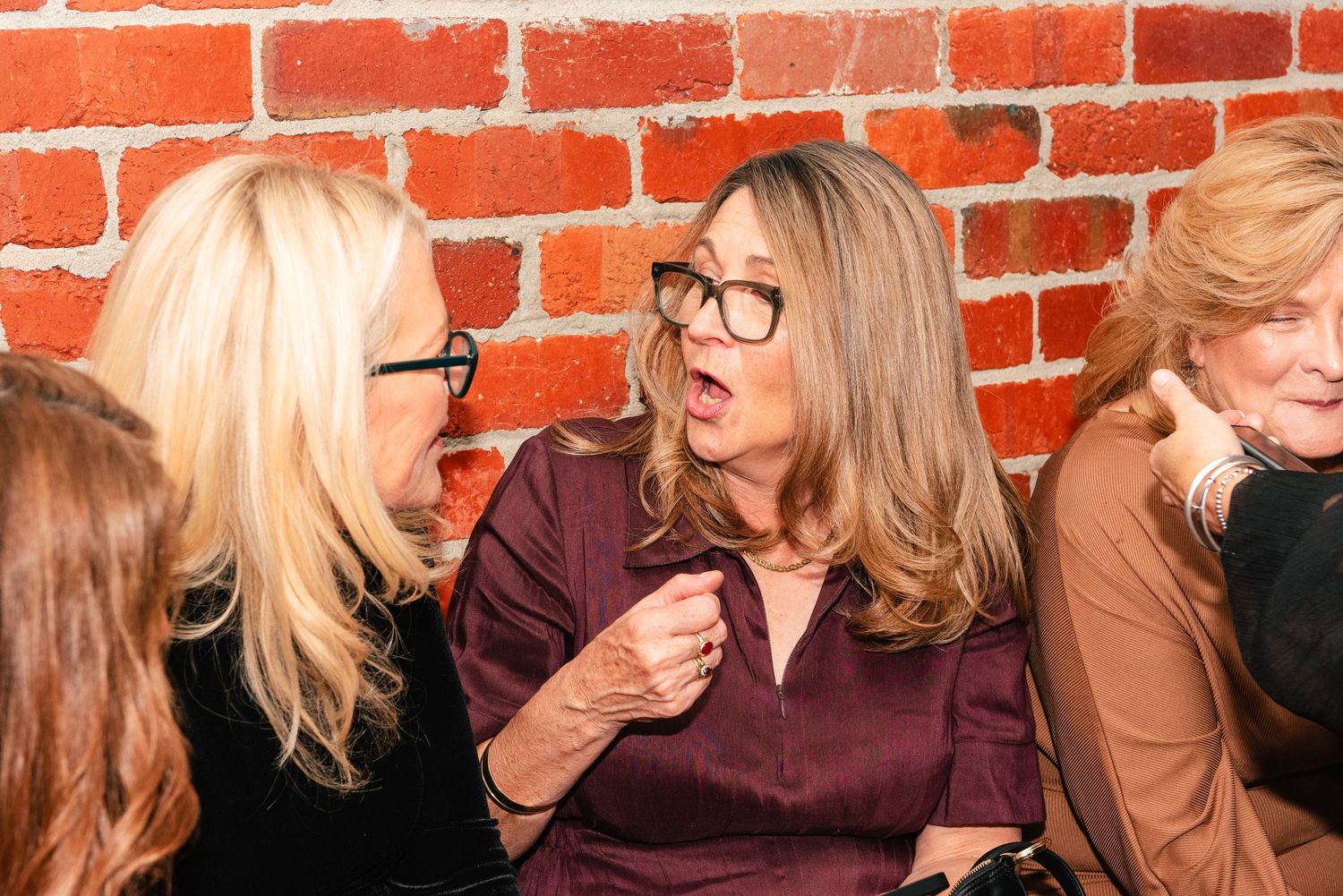 Two women wearing glasses engaged in lively conversation at a social event near a brick wall.