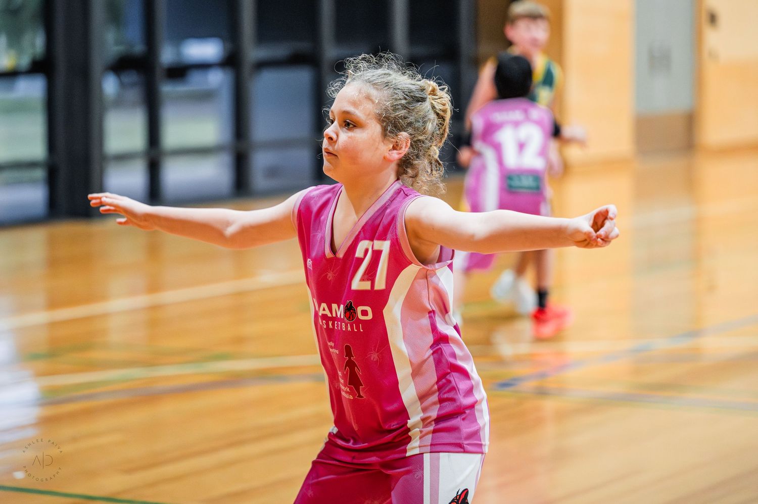 Young girl in pink jersey number 27 playing basketball, arms outstretched on indoor court.