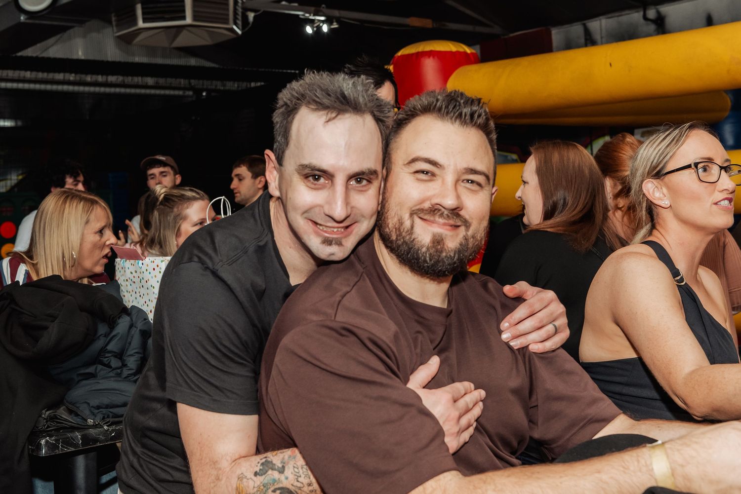 Two smiling men pose together at a lively bar or nightclub event with guests in the background.