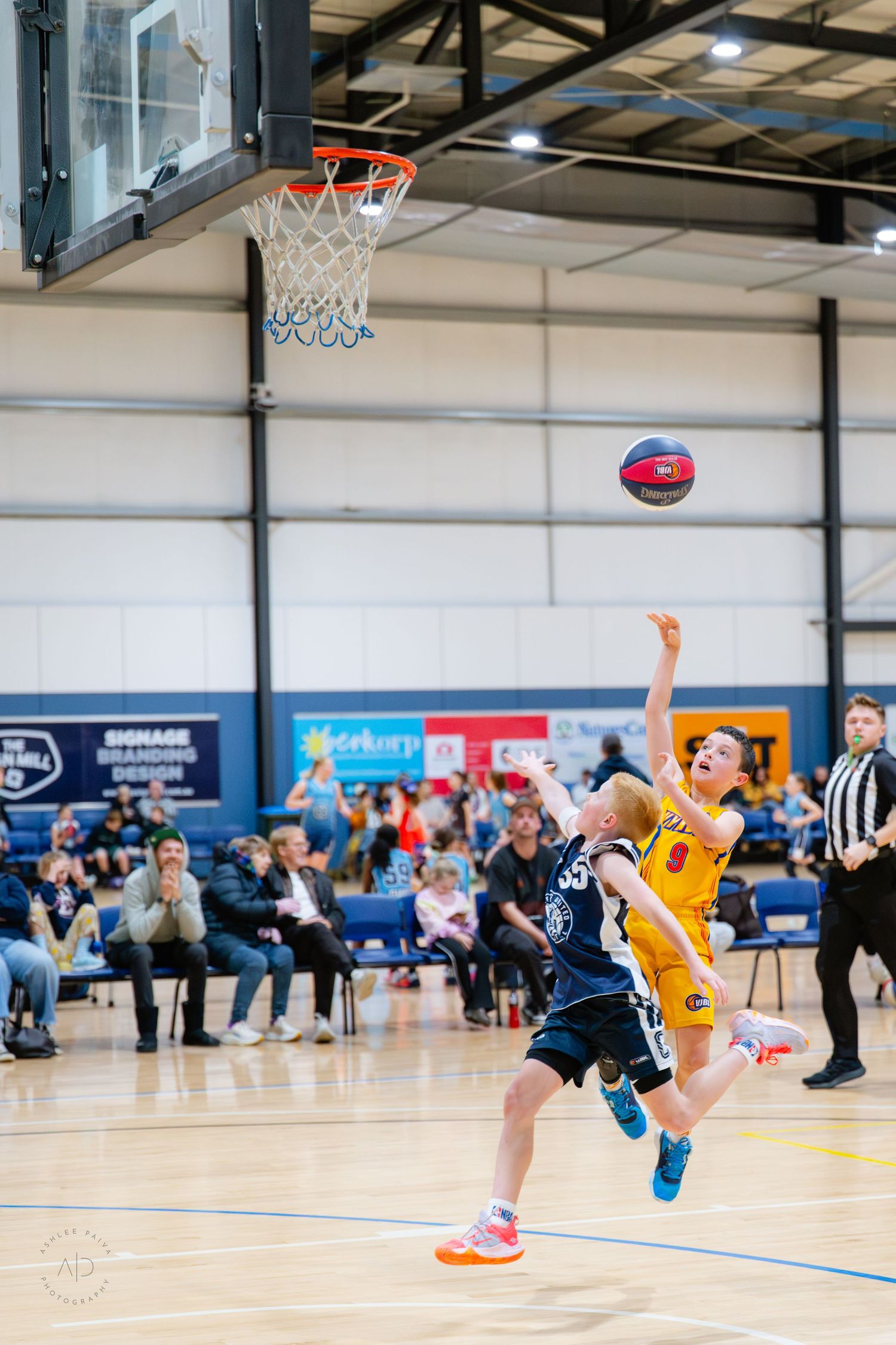 Two young basketball players compete during an indoor game, one in yellow attempting a layup shot toward the hoop.