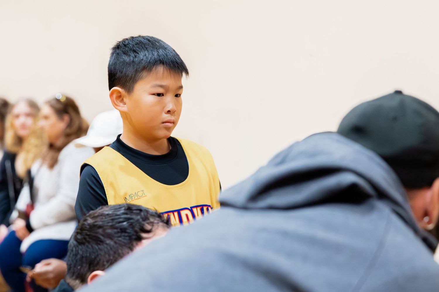 Young boy in yellow basketball jersey stands attentively in a gym during a youth sports event.