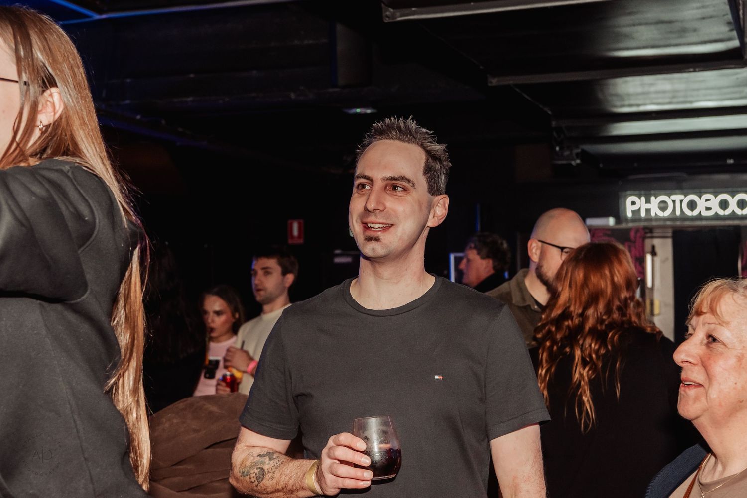 Man holding a drink at a dark venue party, smiling, with a photobooth sign visible in the background.