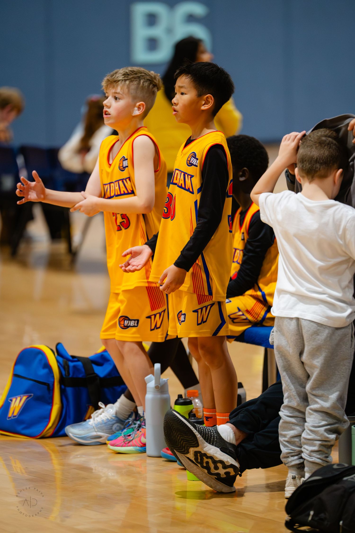 Young basketball players in yellow uniforms stand on the sideline during a youth basketball game in an indoor gym.