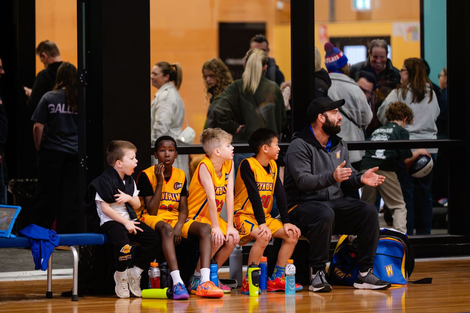 Youth basketball players in gold uniforms sit on a bench with a coach during a game at an indoor court.