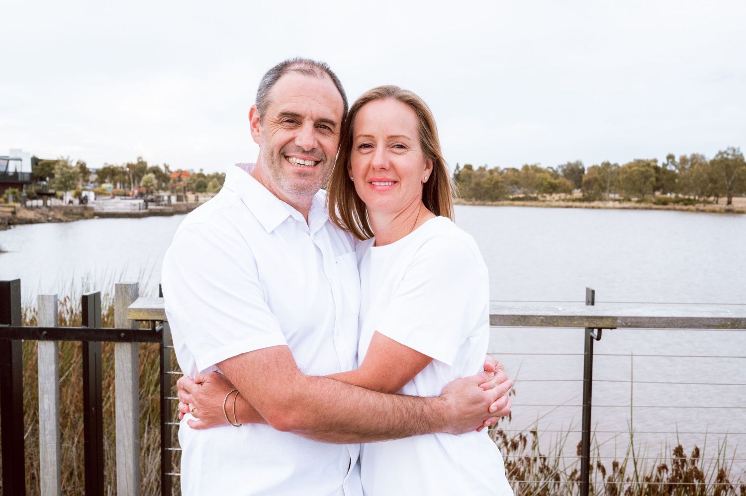 A couple in white shirts embraces while smiling on a waterfront deck.