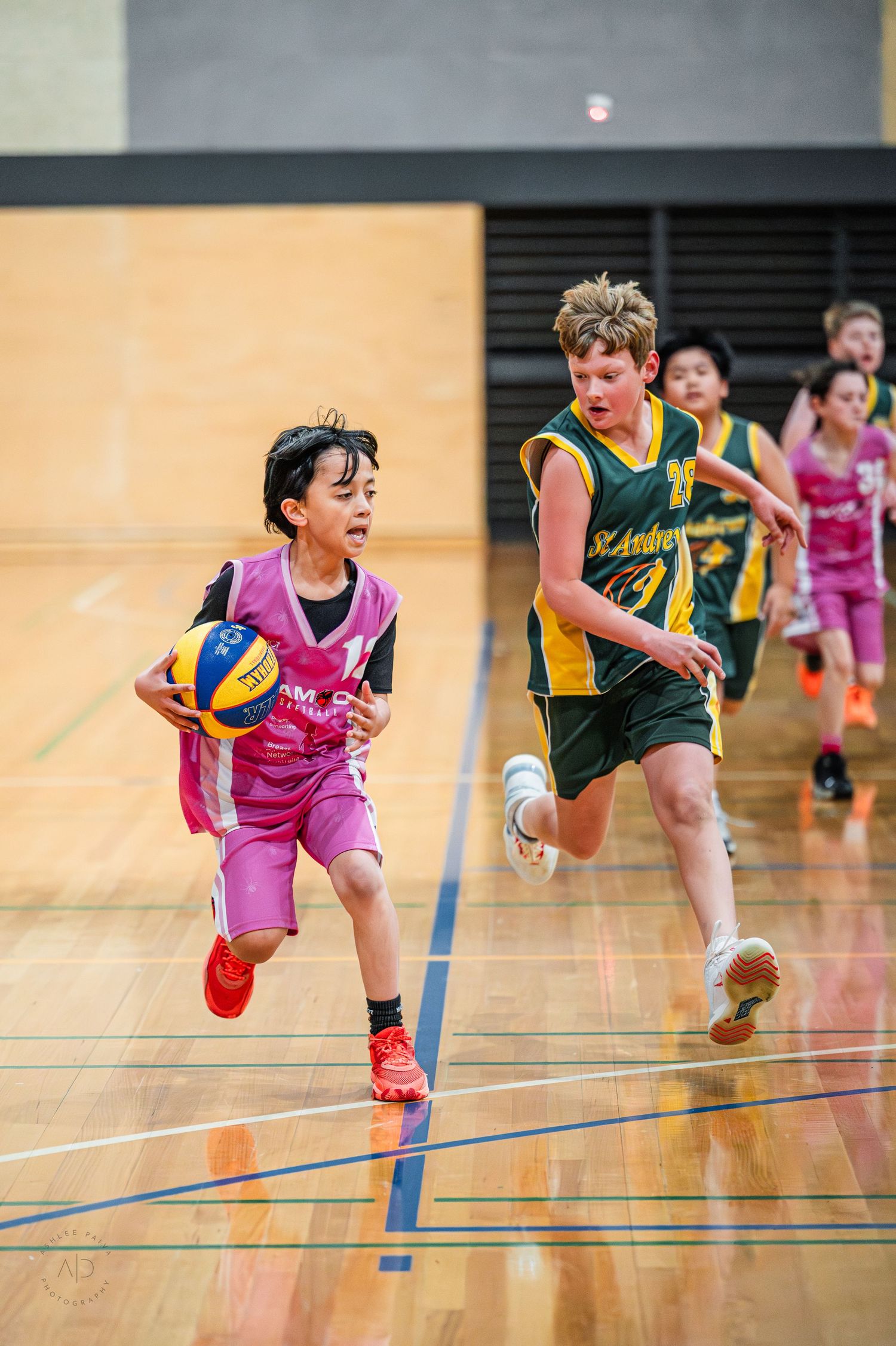 Young boys playing indoor basketball, one in pink uniform dribbling past a defender in green during a youth league game.