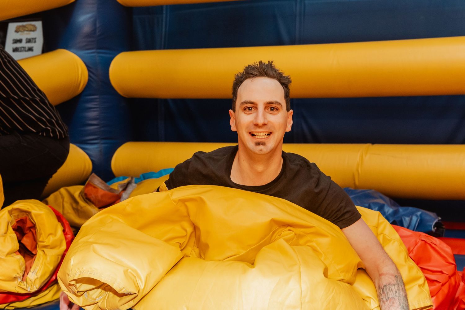 Man smiling inside a colorful bounce house surrounded by large yellow inflatable cushions.