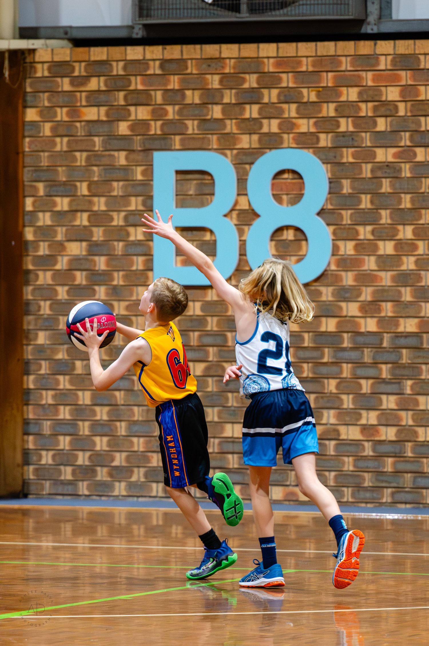 Two young kids playing basketball indoors, one in yellow jersey holding ball while defender in white reaches up to block.