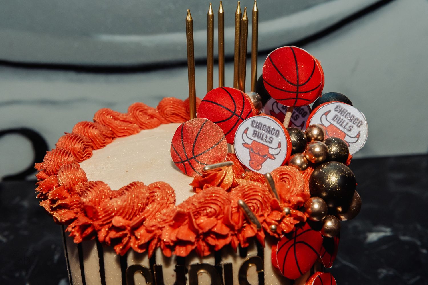 Chicago Bulls themed birthday cake with basketball decorations, orange rosette frosting, gold candles, and team logo toppers.