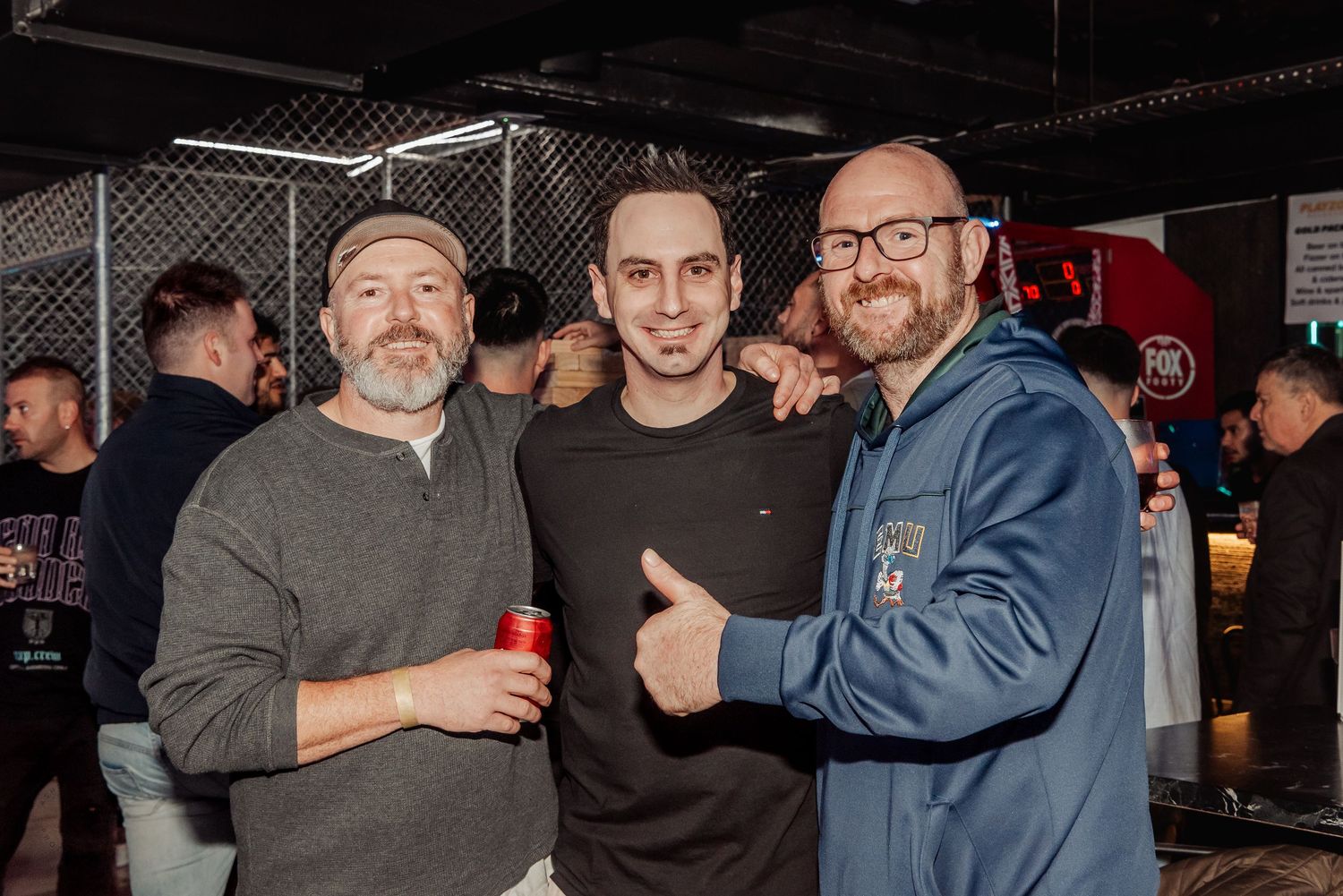 Three men pose together smiling at a social event, one holding a red drink can in a dimly lit venue.