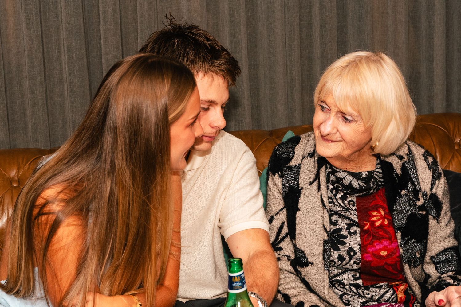 Three people sitting on a brown leather sofa, smiling and chatting at a social gathering.