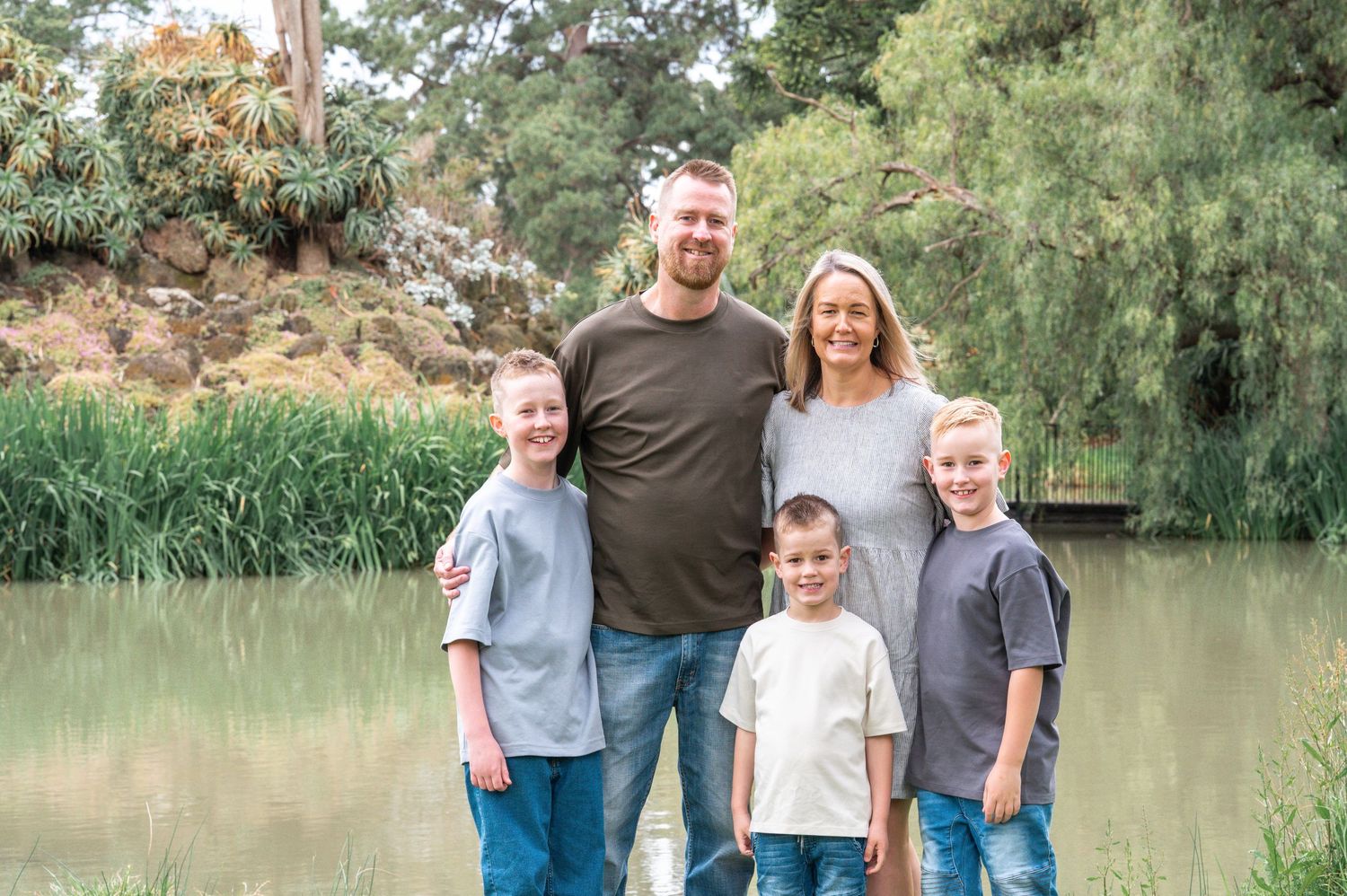 A family of five poses together in front of a serene pond with greenery in the background.