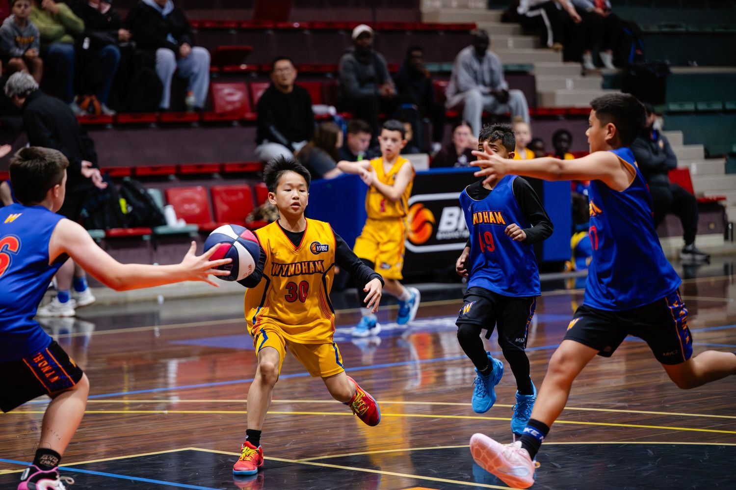 Youth basketball players compete in an indoor court game, with a player in yellow dribbling against blue-uniformed defenders.