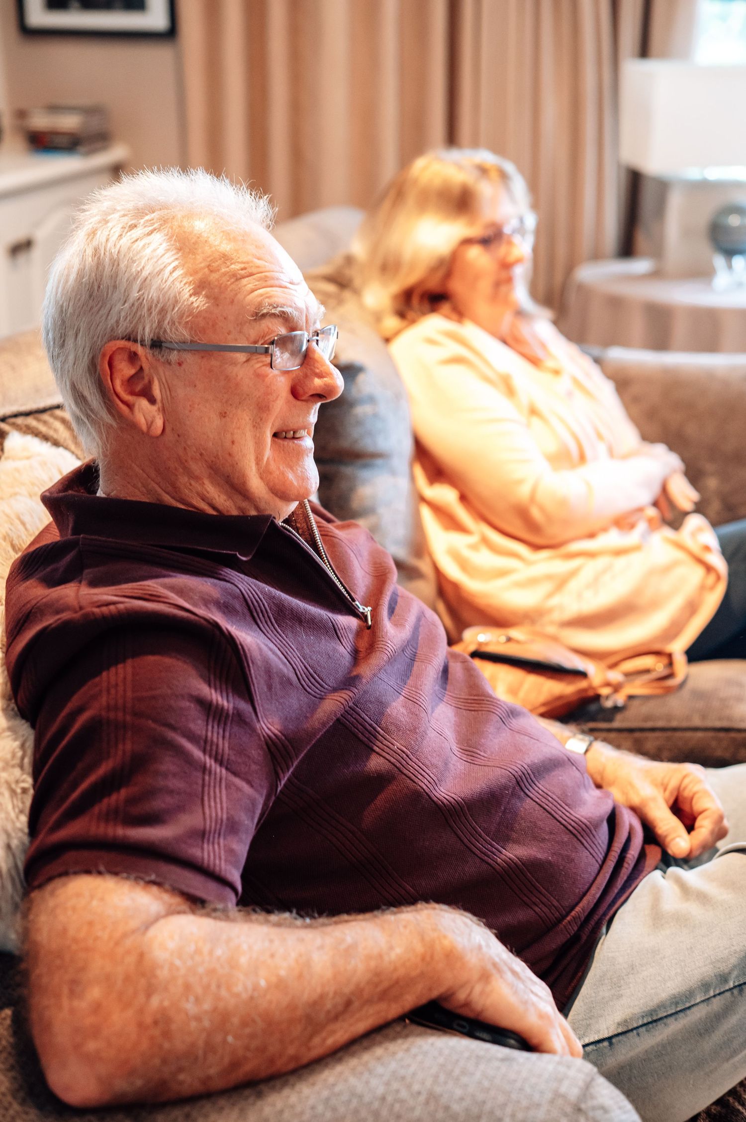 Senior couple relaxing on a couch in a cozy living room, man in maroon polo shirt wearing glasses.