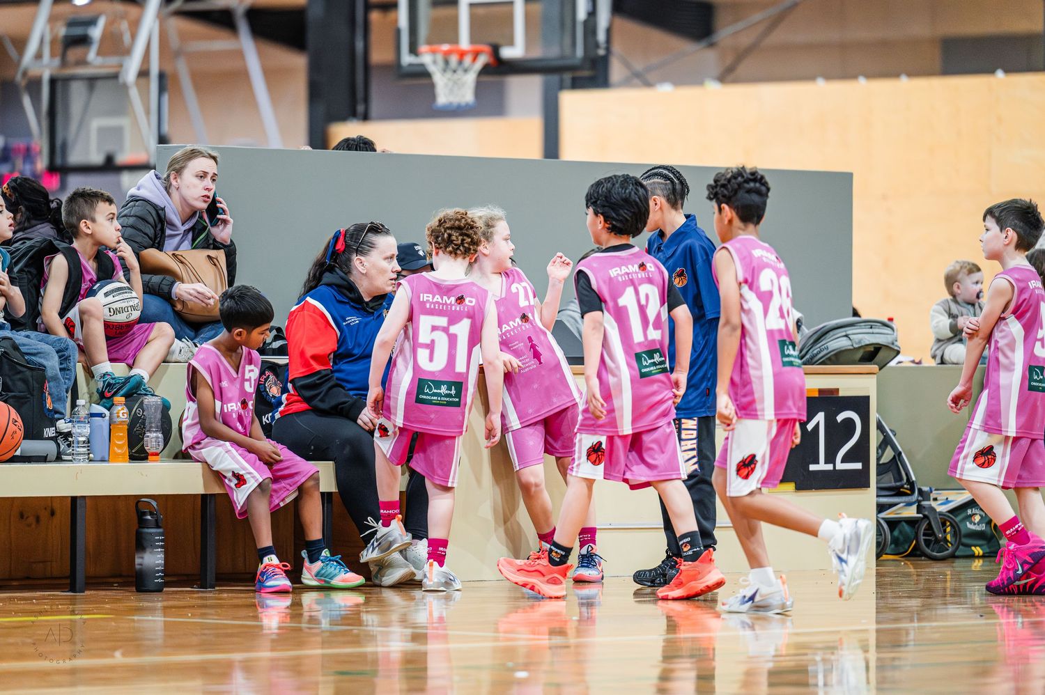 Youth basketball players in pink uniforms huddle near the bench during a junior league game indoors.