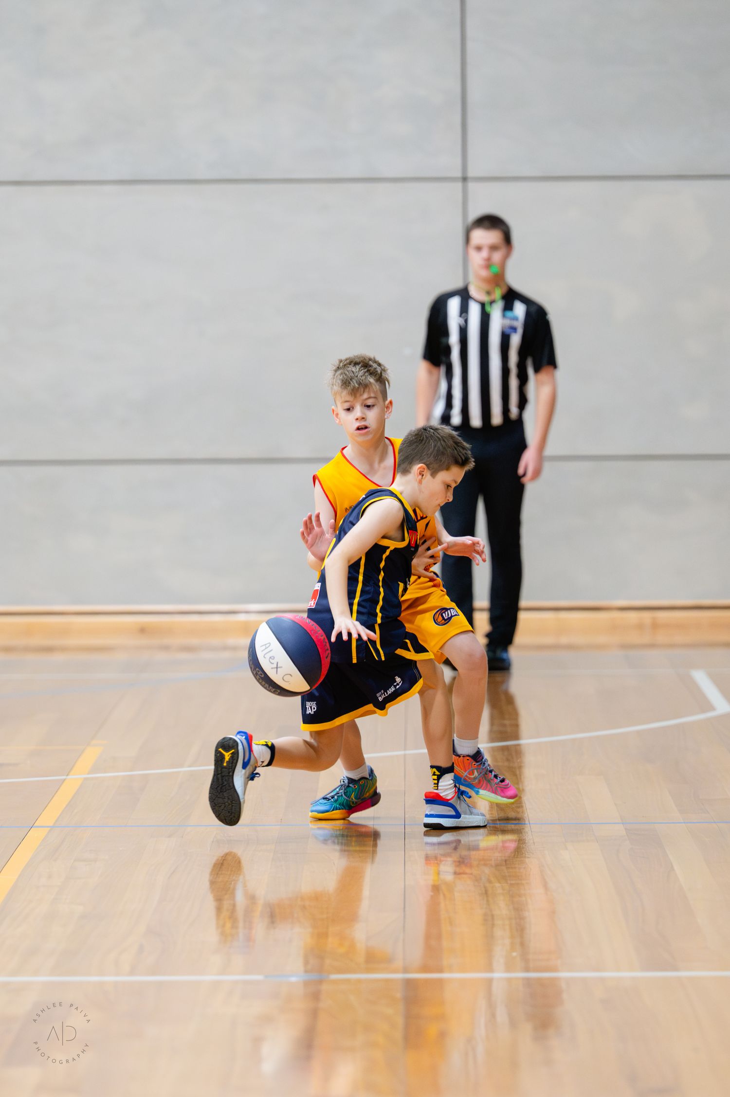Two young boys in yellow uniforms compete for a basketball on an indoor court with a referee watching in the background.