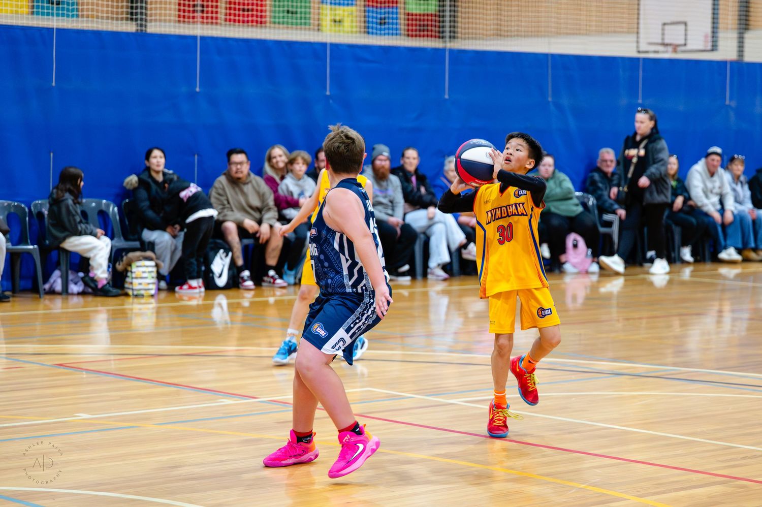 Two youth basketball players compete on an indoor court, one in yellow shooting while defender in blue watches.