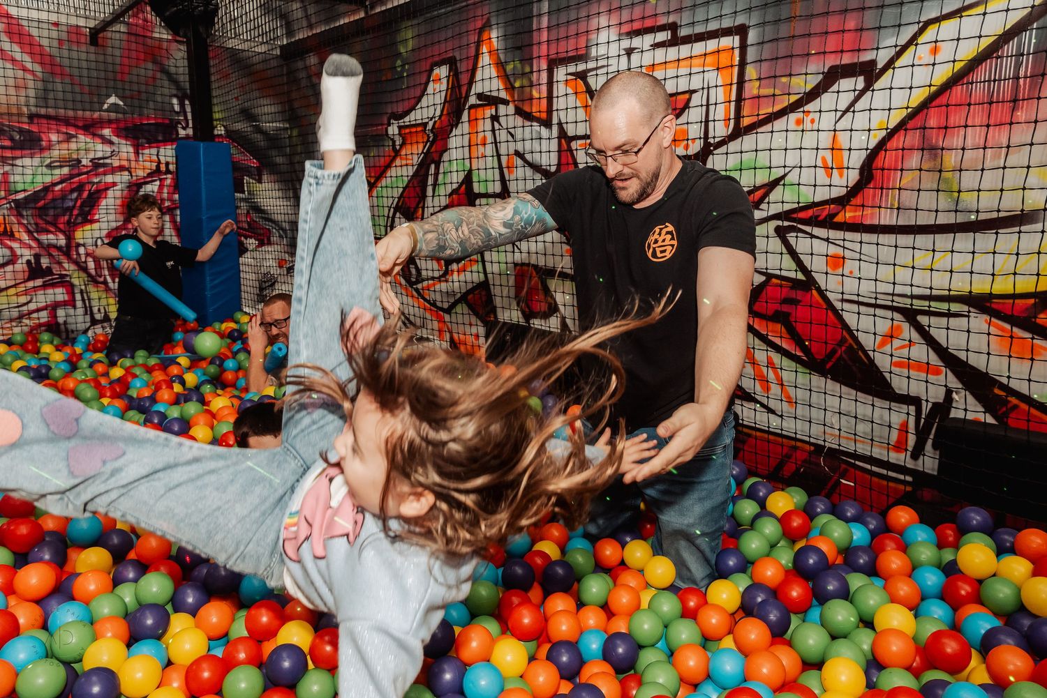 Man and child playing in colorful ball pit at indoor playground with graffiti wall backdrop.