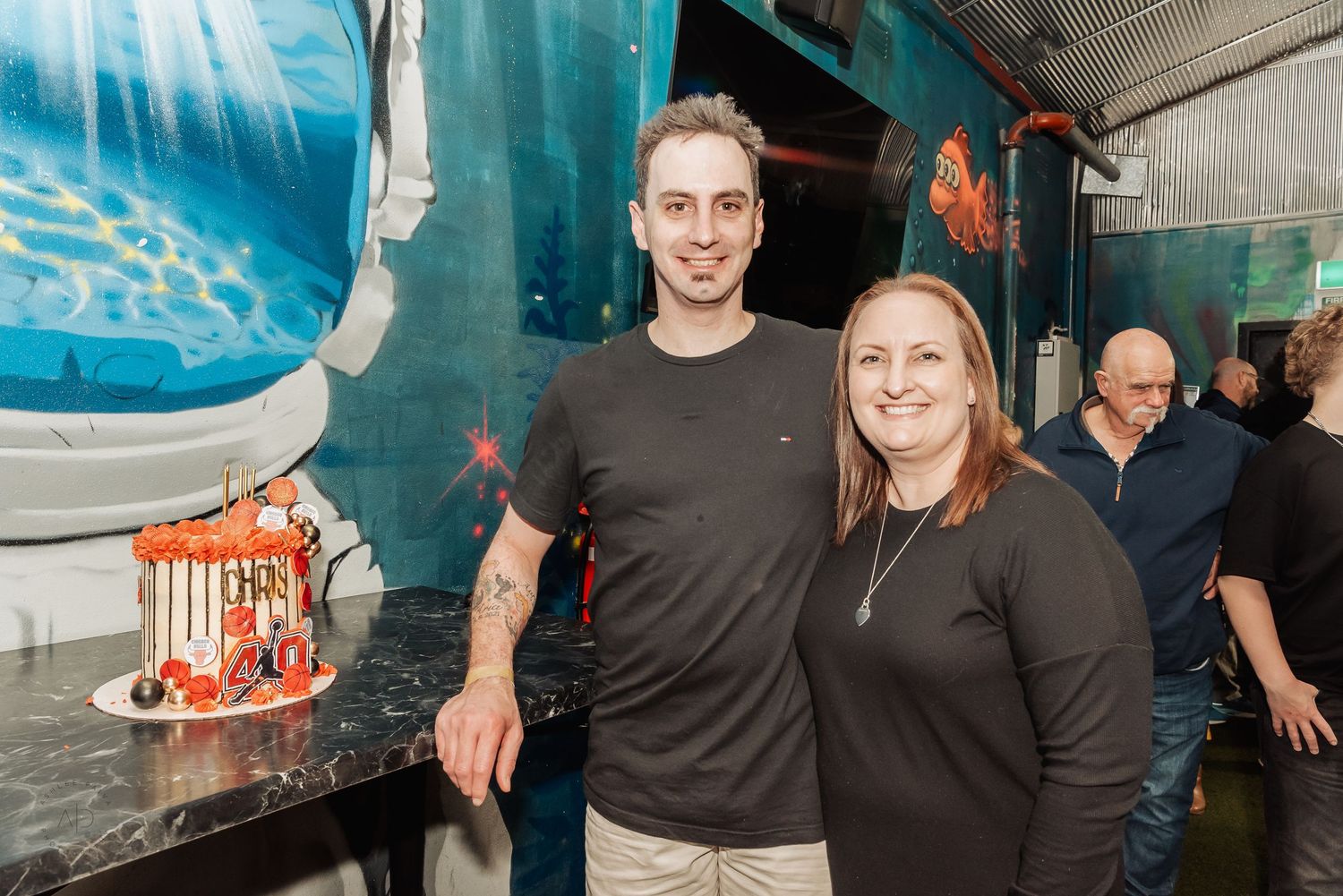 Two people smiling next to a decorated birthday cake at a vibrant indoor party venue.