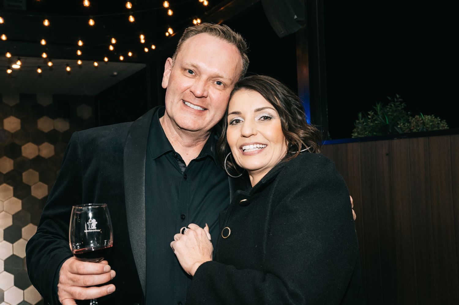 Couple smiling together at nighttime event with string lights in background.