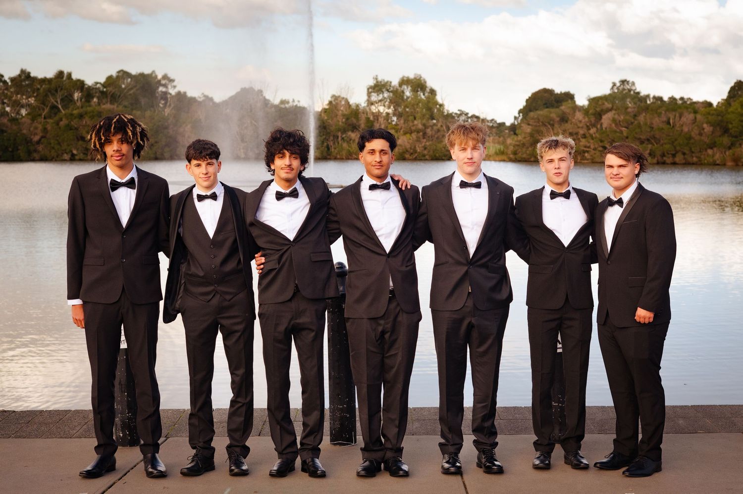 Seven young men in black tuxedos with bow ties pose together outdoors near a fountain for a formal event.