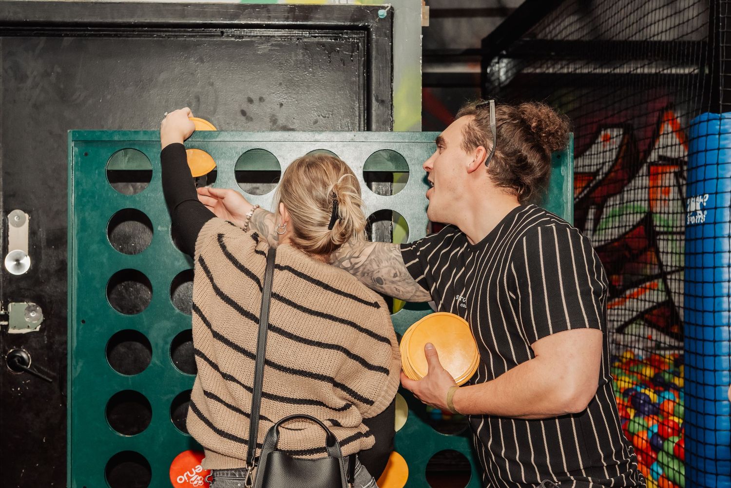 Two people playing a giant Connect Four game at an indoor play center with colorful ball pit in background.