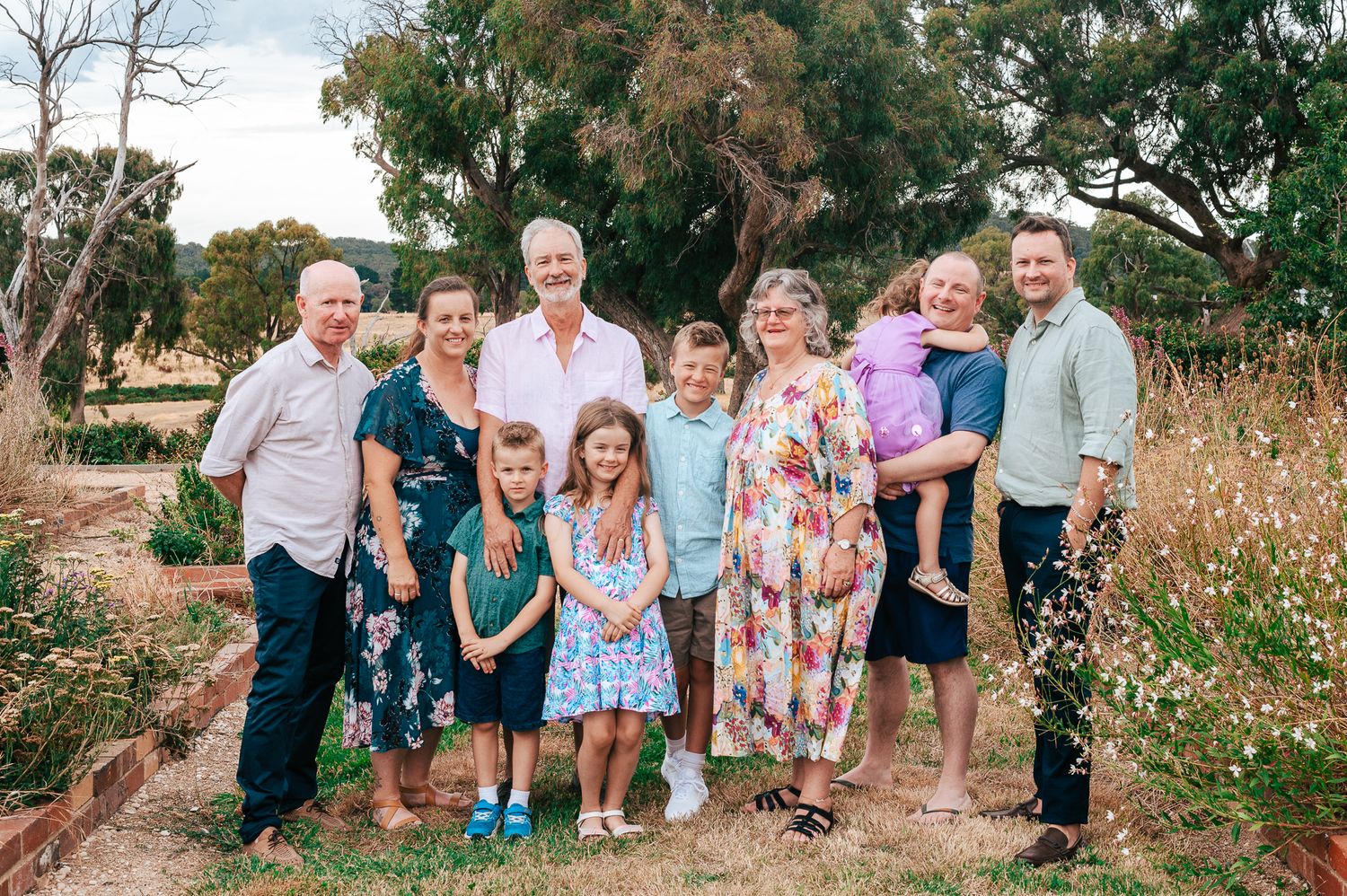 A group of people stand together in a natural outdoor setting wearing casual clothing and floral dresses.