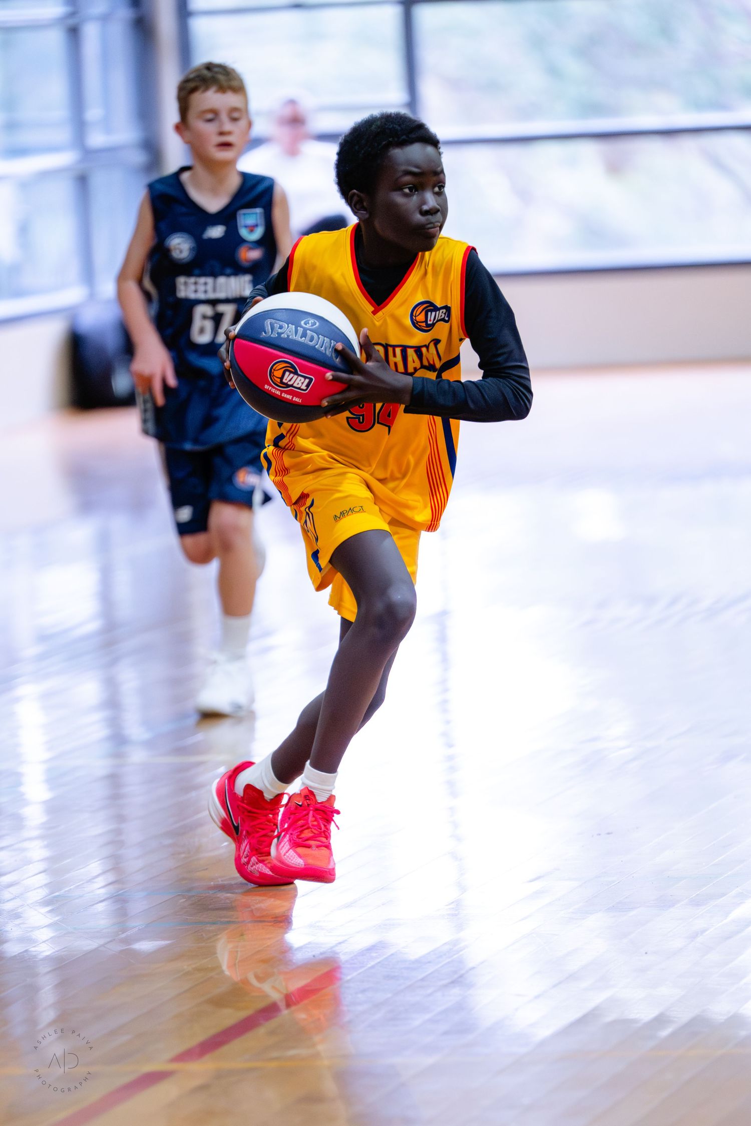 Young basketball player in yellow uniform dribbles ball during indoor youth game, defender in blue Geelong jersey behind.