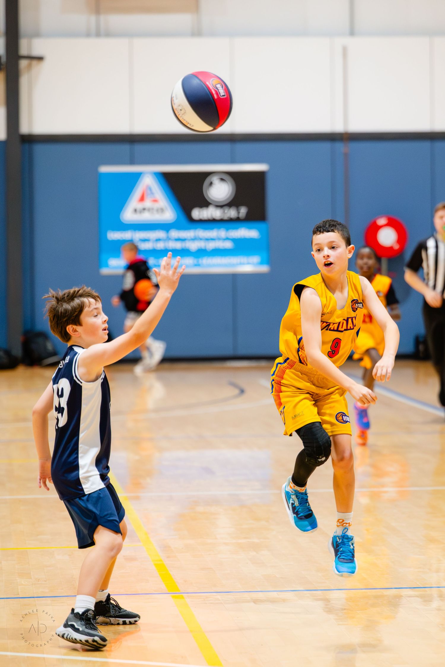 Two boys playing youth basketball indoors, one in yellow uniform jumping to pass the ball to teammate in navy uniform.