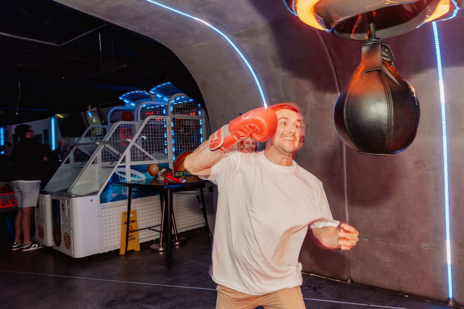 Man in white t-shirt throwing a punch at a speed bag in an arcade game room wearing orange boxing gloves.