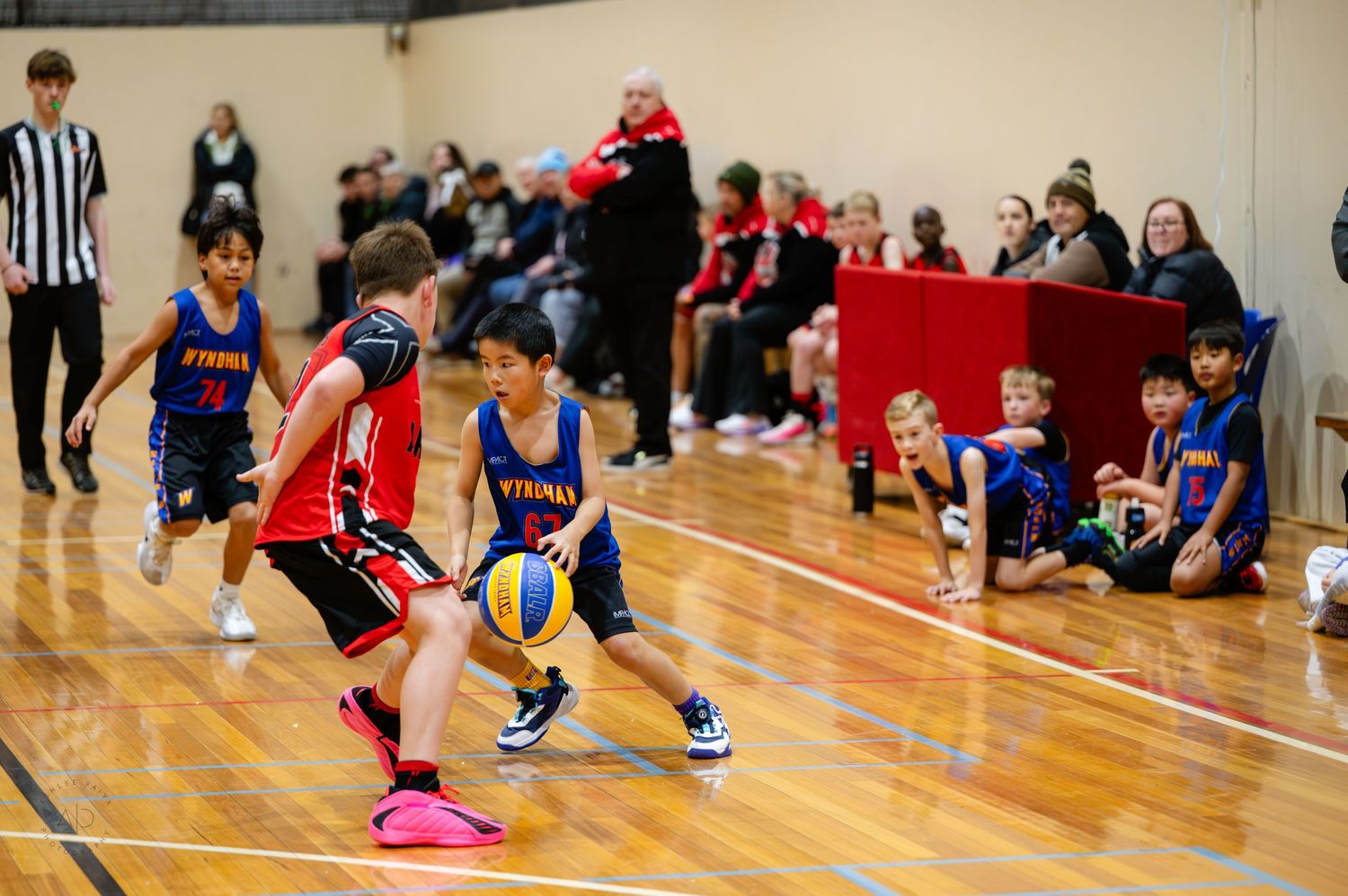 Young boys playing basketball in an indoor court during a youth league game, with spectators watching from the sideline.