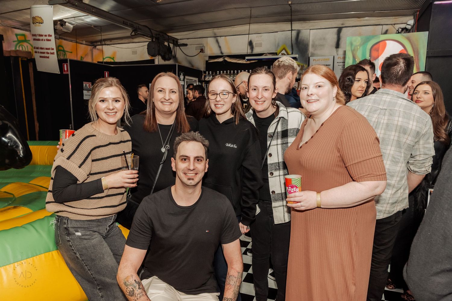 Group of friends posing together at an indoor event venue with colorful decorations in the background.