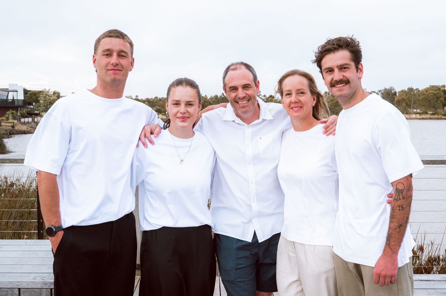 A group of five people wearing white tops poses together on a dock by the water.