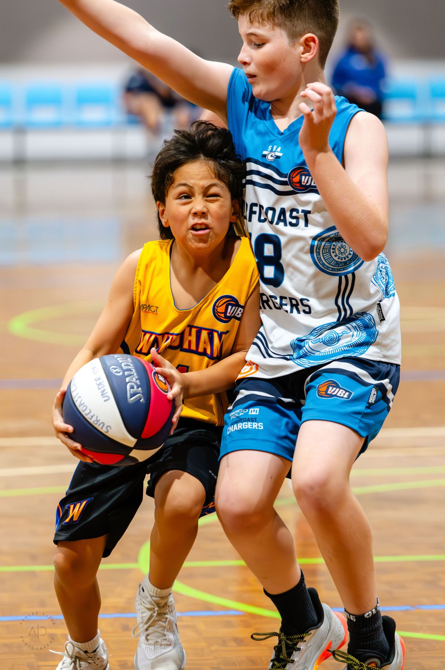 Two youth basketball players compete on court, one in yellow driving past a defender in blue Gulf Coast Chargers uniform.