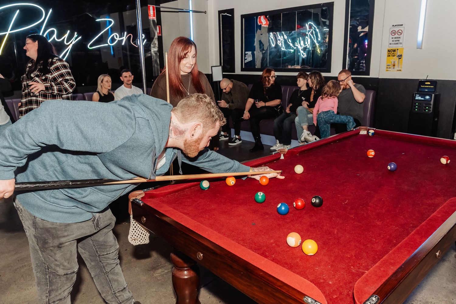 A player leans over a red pool table taking a shot in a lively Play Zone bar with spectators watching in the background.