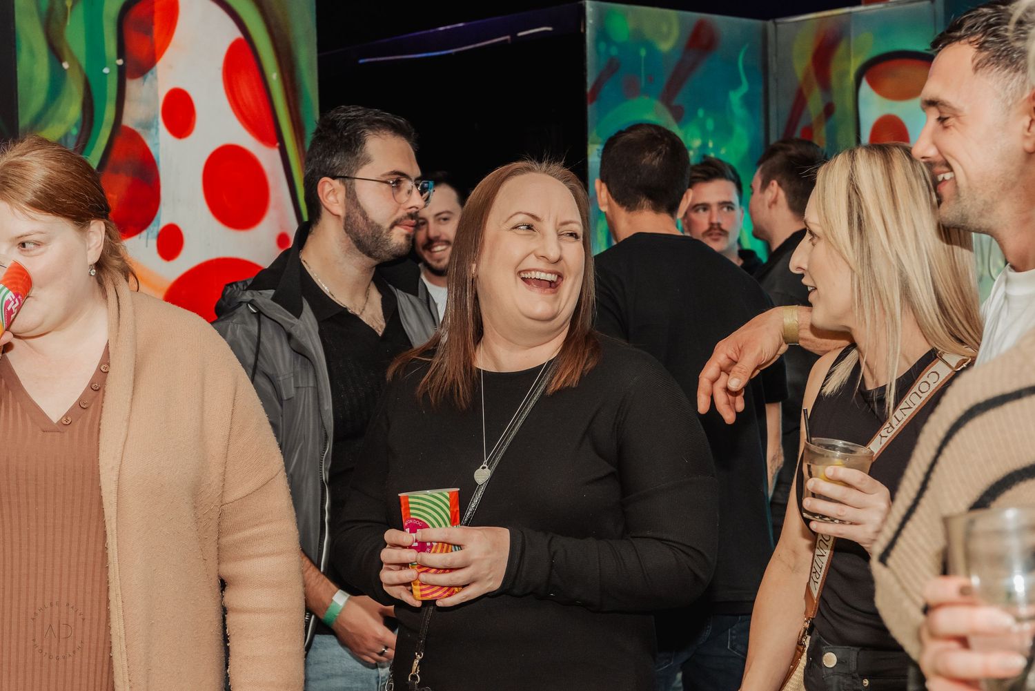 Group of people socializing at a colorful indoor party event, smiling and holding drinks.