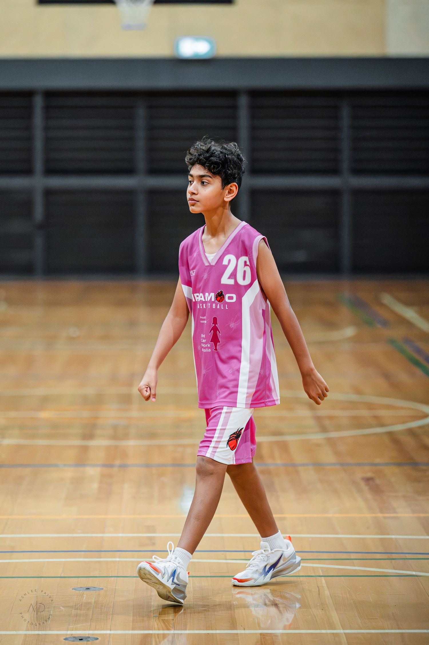 Young basketball player wearing pink number 26 jersey walking on indoor court during game.