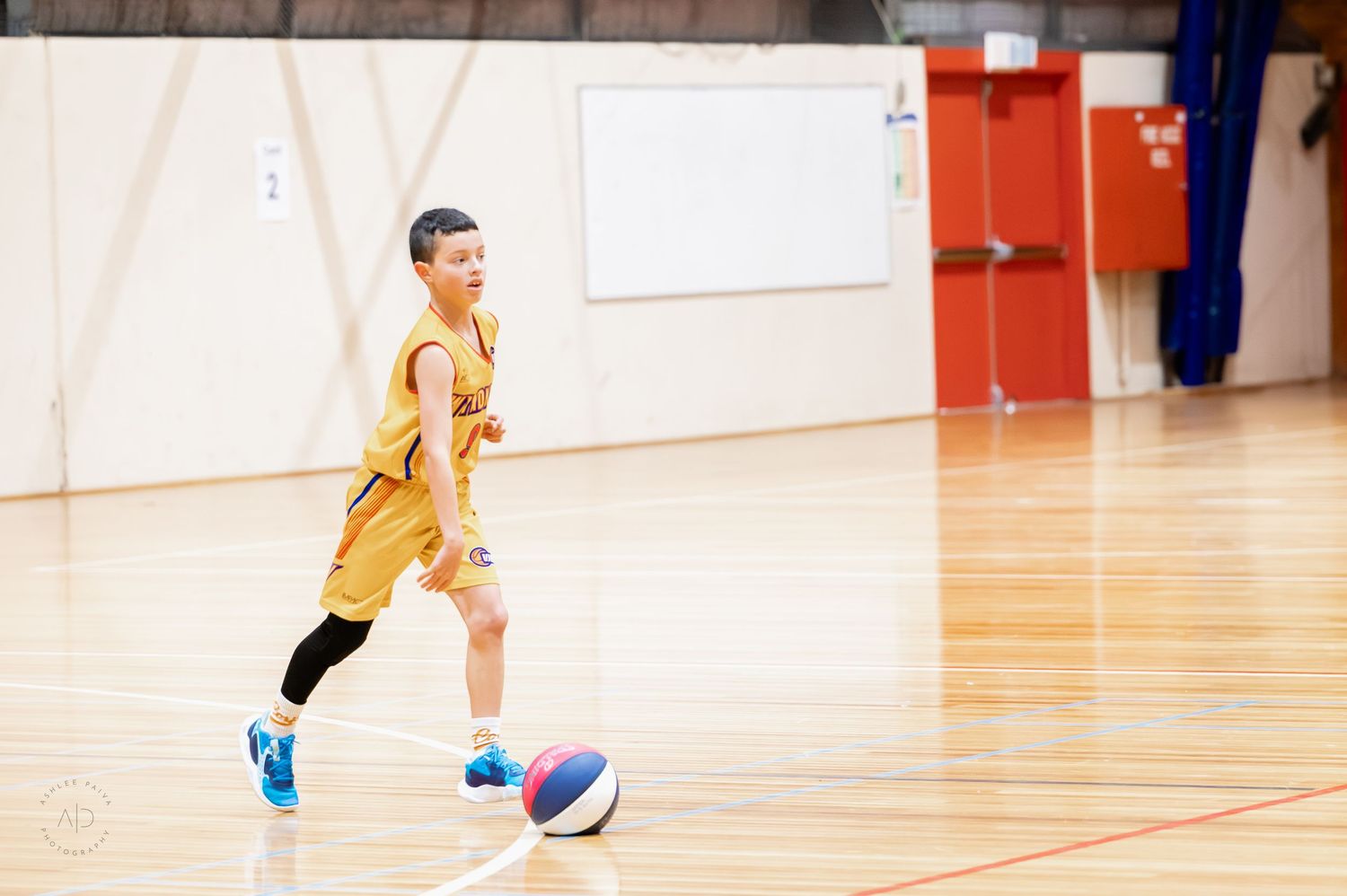 Young boy in yellow basketball uniform dribbling a colorful ball on an indoor hardwood court.