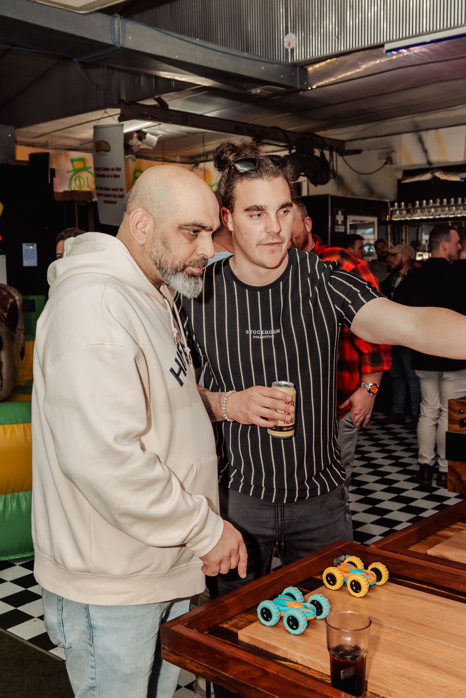 Two men pose together in a bar with checkered floors, one holding a beer, with colorful toy trucks on a wooden table.