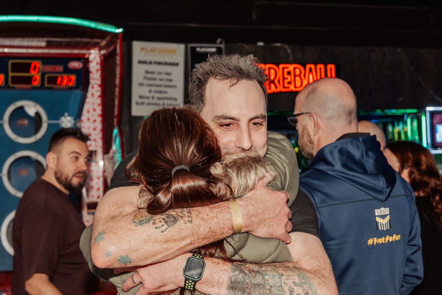 Two people share a warm hug at an arcade bar with neon signs and Fireball branding in the background.