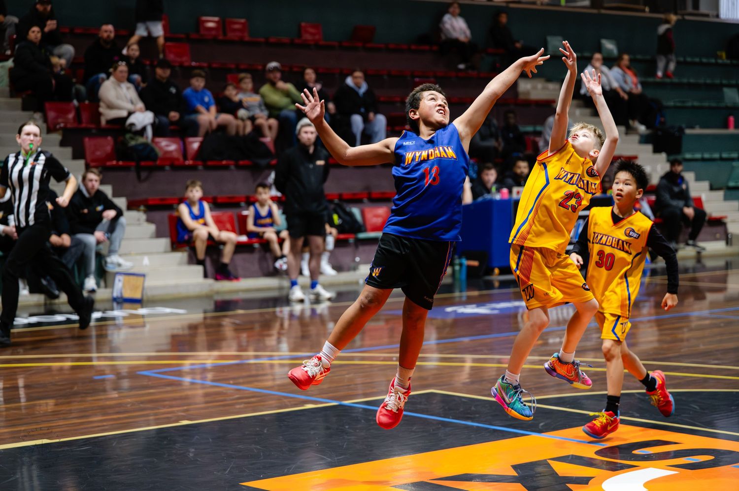 Basketball player in blue jersey leaps for a shot during an indoor game, defenders in yellow uniforms close behind.