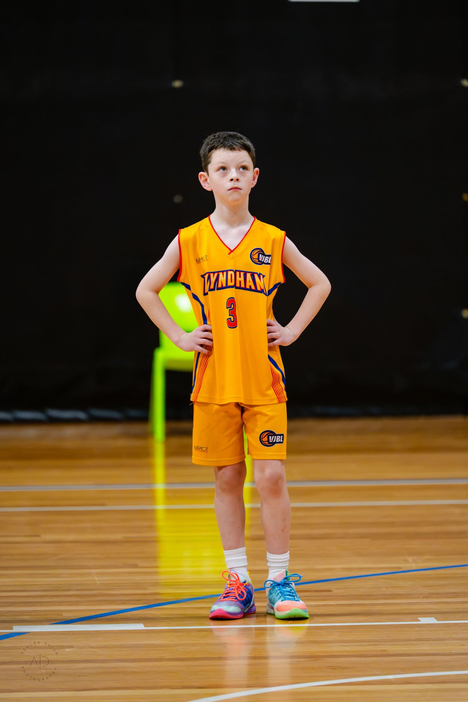 Young boy in orange Wyndham basketball uniform number 3 stands confidently on indoor court with hands on hips.