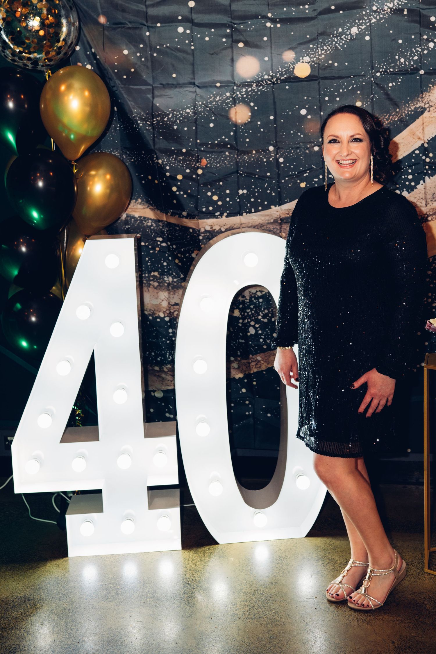 Woman in black dress poses next to illuminated number 40 sign with gold balloons in background.