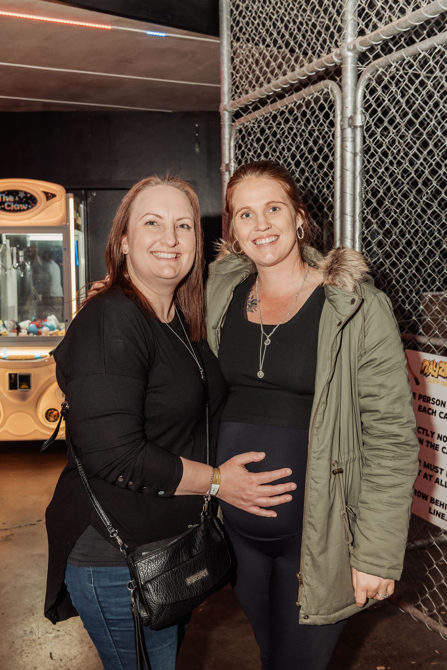 Two women smiling at a carnival, one visibly pregnant, standing near a claw machine arcade game at night.
