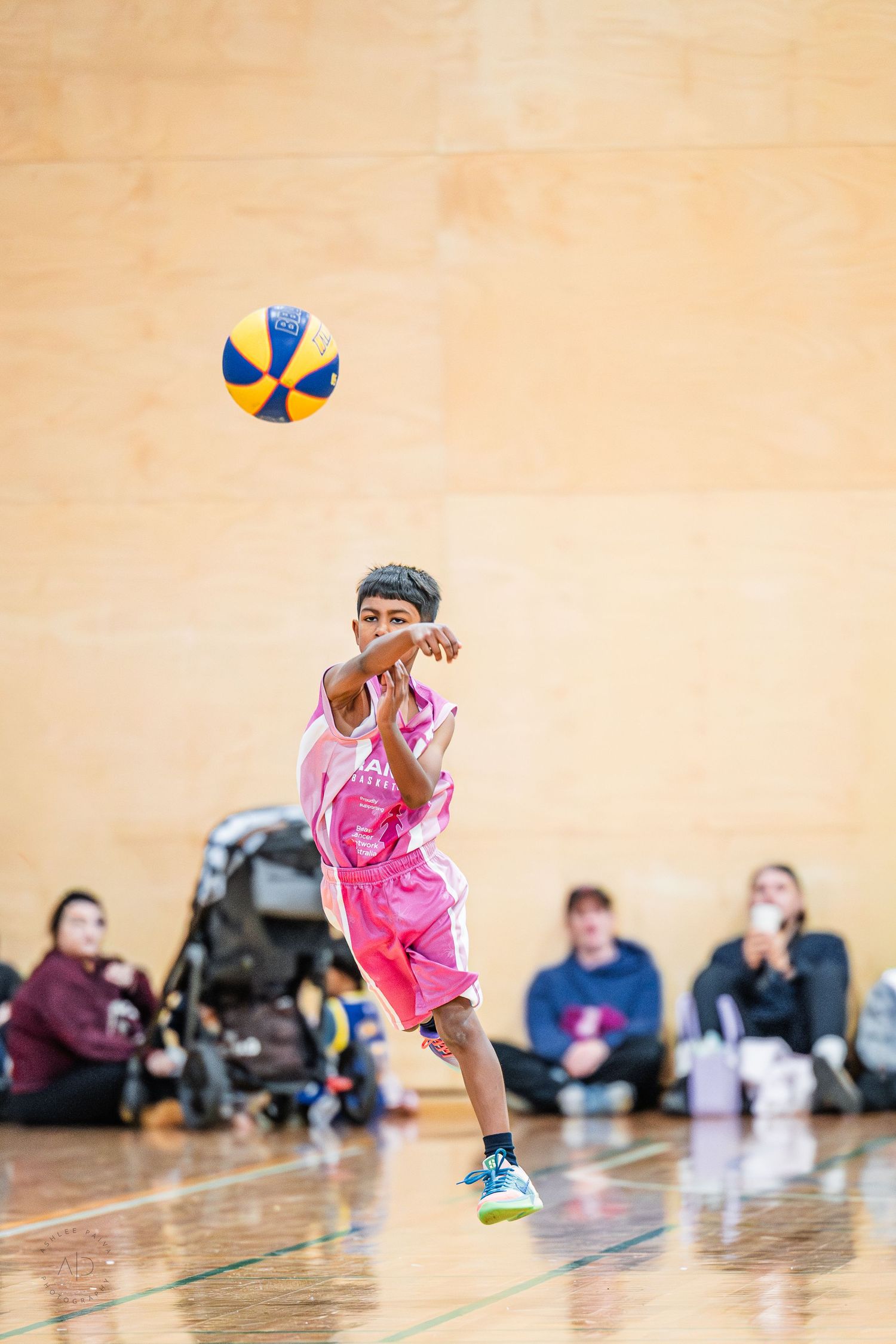 Young boy in pink basketball uniform jumping to pass a yellow and blue ball during an indoor youth basketball game.